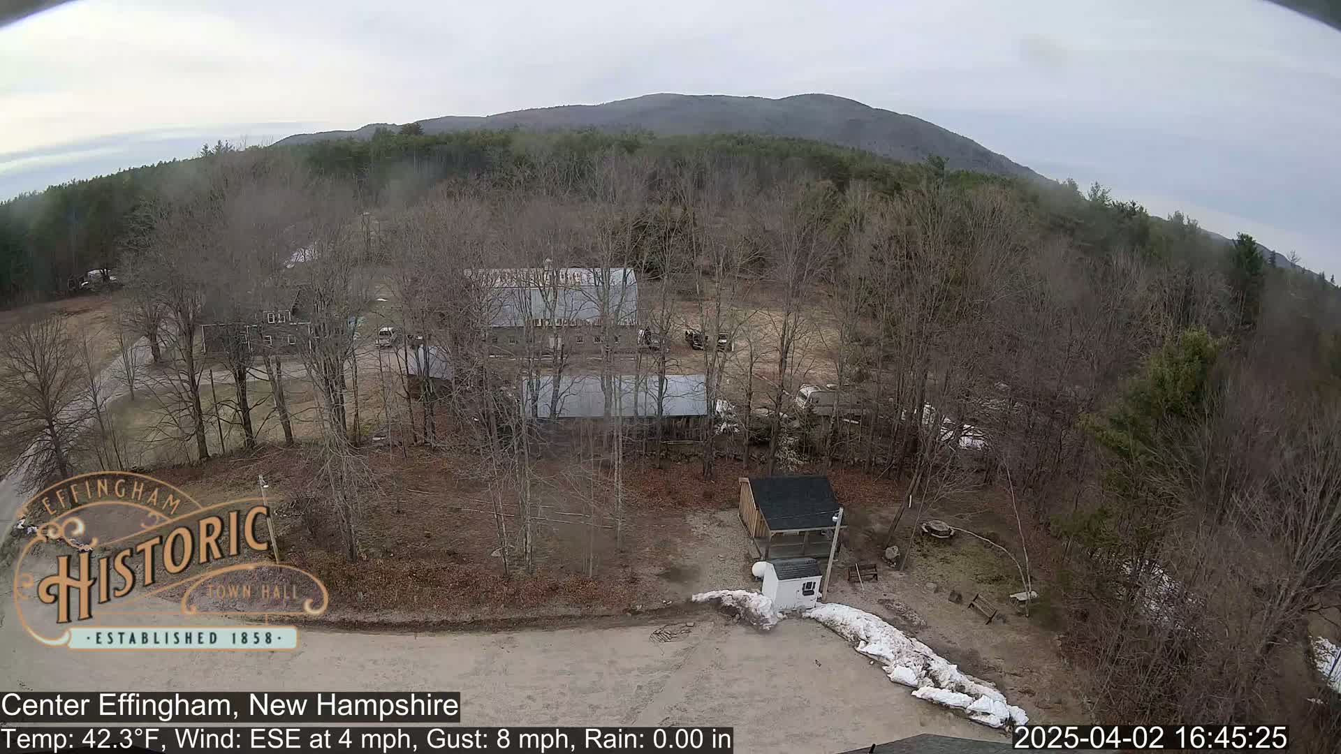 An aerial view shows a rural area with several buildings, bare trees, and a small amount of snow on the ground under overcast skies.