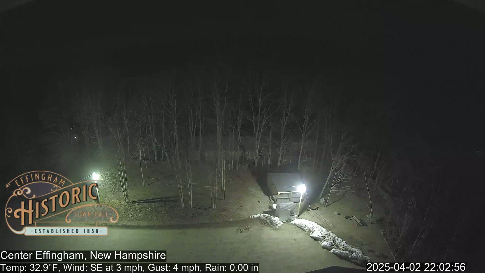 A nighttime view of a sparsely lit area with bare trees and some snow on the ground, under a clear, dark sky.