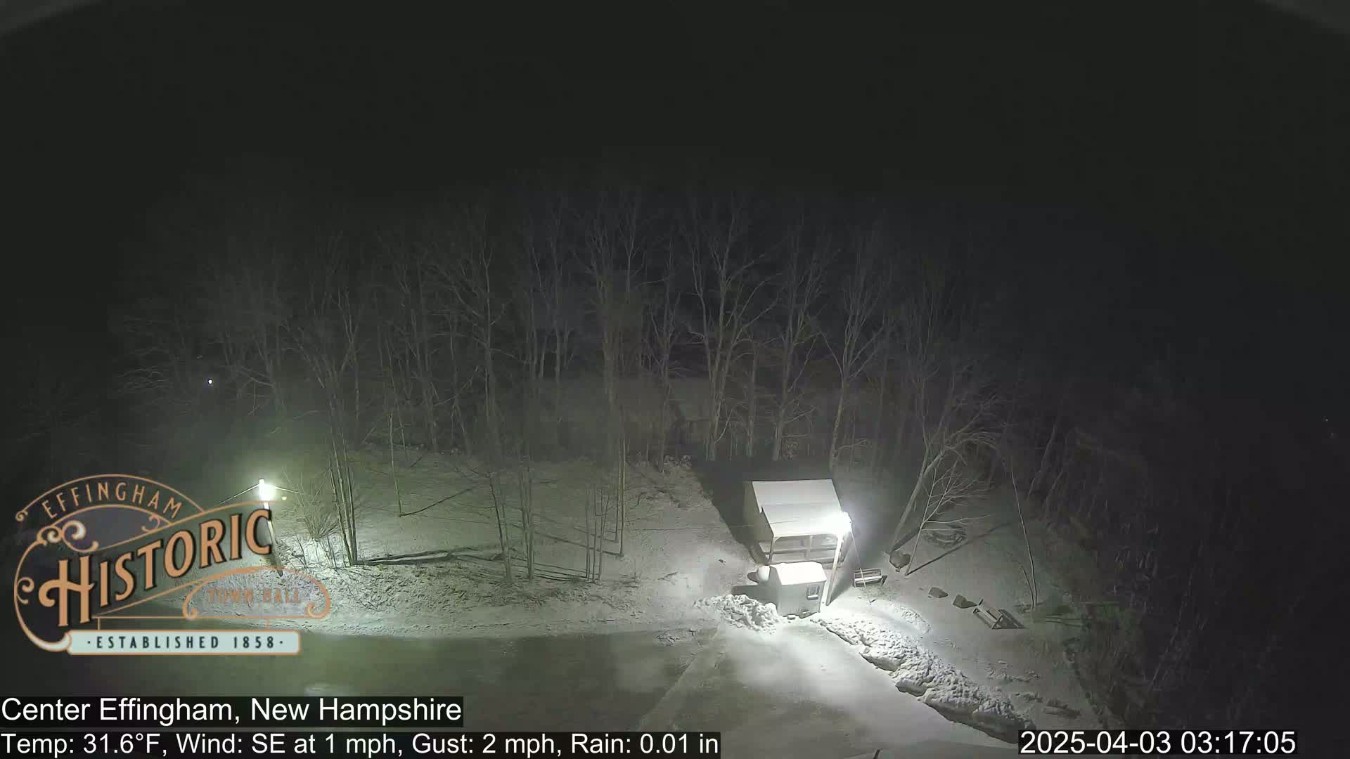 A nighttime, snowy scene shows a small structure illuminated by lights, surrounded by bare trees and a snow-covered ground under a dark sky.
