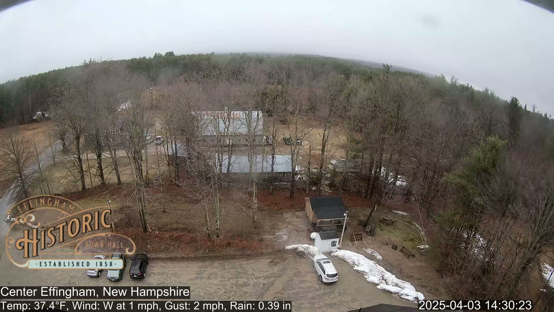An aerial view shows a rural area with several buildings, cars, and patches of snow under an overcast sky with light rain.
