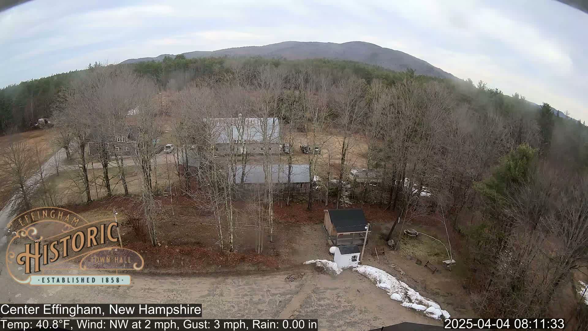 An aerial view shows several buildings in a clearing surrounded by bare, deciduous trees and a distant mountain range under an overcast sky.