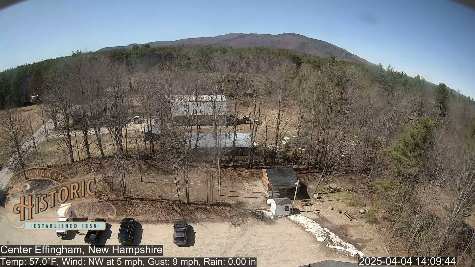 A high-angle view of a rural area with several buildings, bare trees, and a few parked cars under a clear, sunny sky.