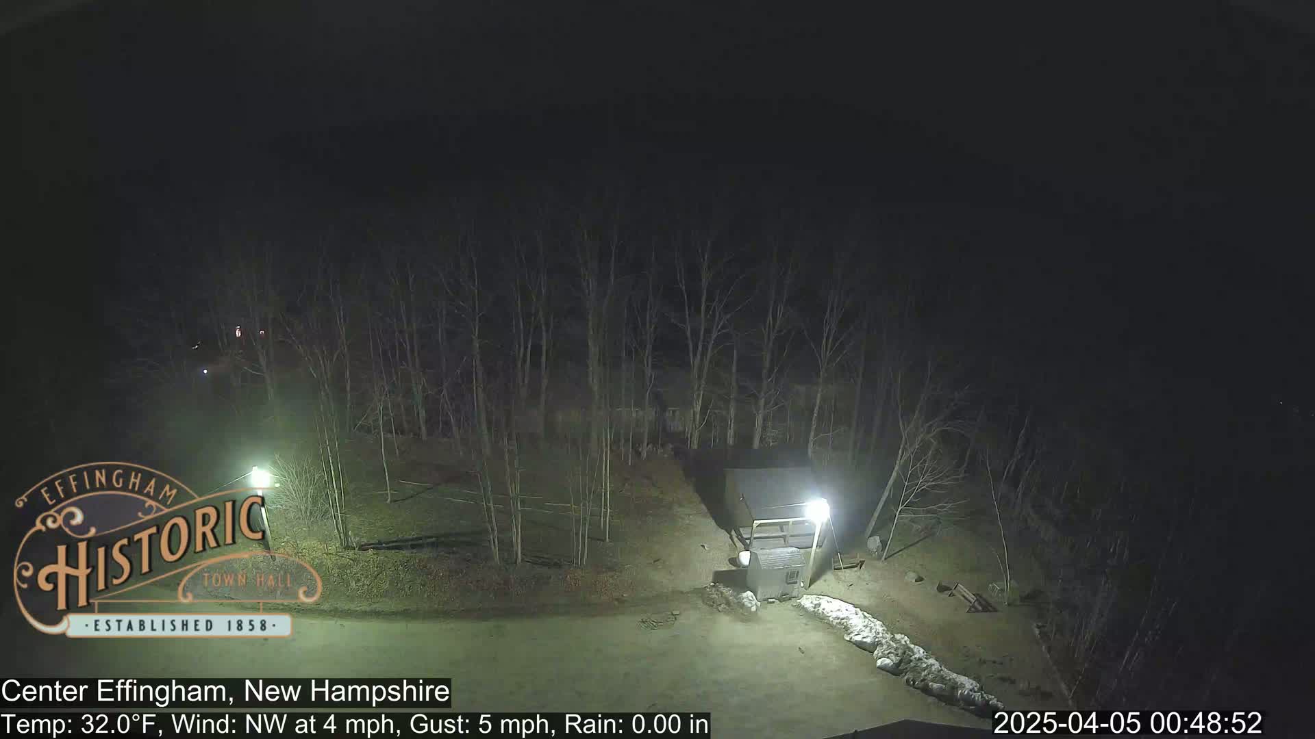 A nighttime view of a sparsely populated area with bare trees, a small building, and a pile of what appears to be snow or ice, under a dark, clear sky; a northwest wind is blowing at 4 mph with gusts up to 5 mph.