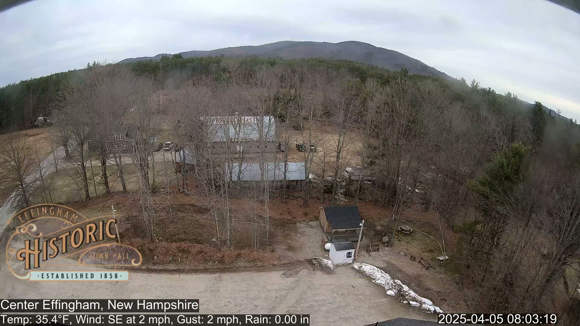 An aerial view of a rural area with several buildings, bare trees, and a small amount of snow on the ground under an overcast sky.