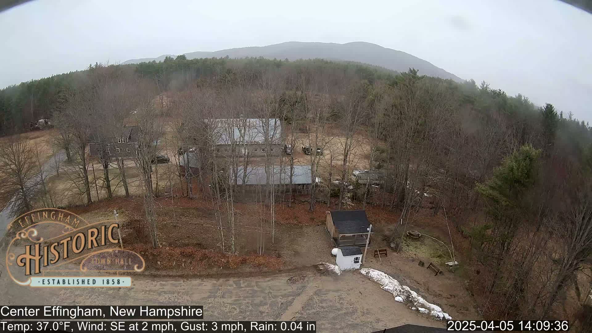 A high-angle view shows several buildings, a mostly bare winter landscape, and light rain under an overcast sky.