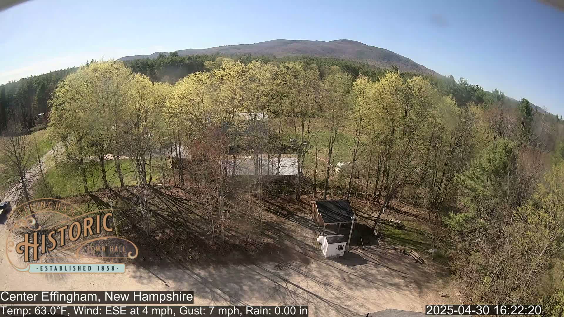 An aerial view shows a small building and a grassy area surrounded by budding trees under a clear sky, with a mountain in the distance.