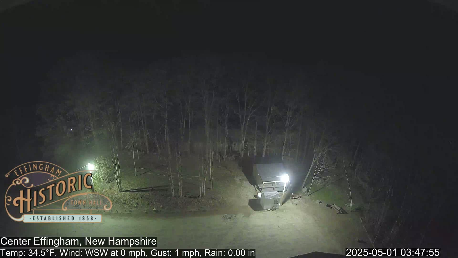 A nighttime view shows a small building in a lightly wooded area under a dark, clear sky.