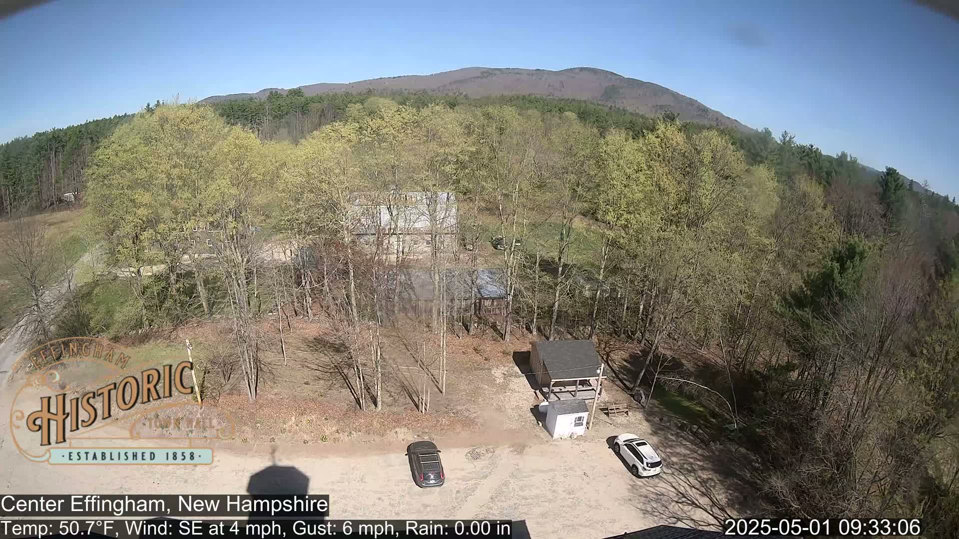 A high-angle, sunny view of a parking lot with two cars, several buildings, and a mostly leafed-out forest extending to distant mountains.