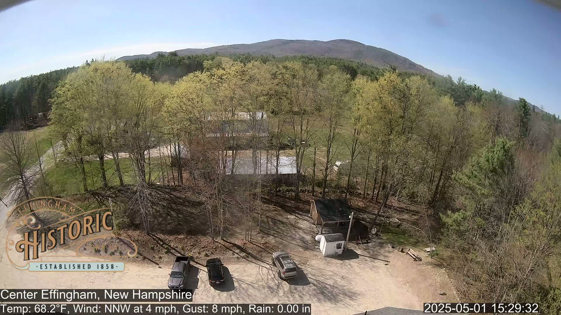 An aerial view shows a parking lot with several cars next to a cluster of buildings surrounded by deciduous trees, a mountain in the background under a clear sky.