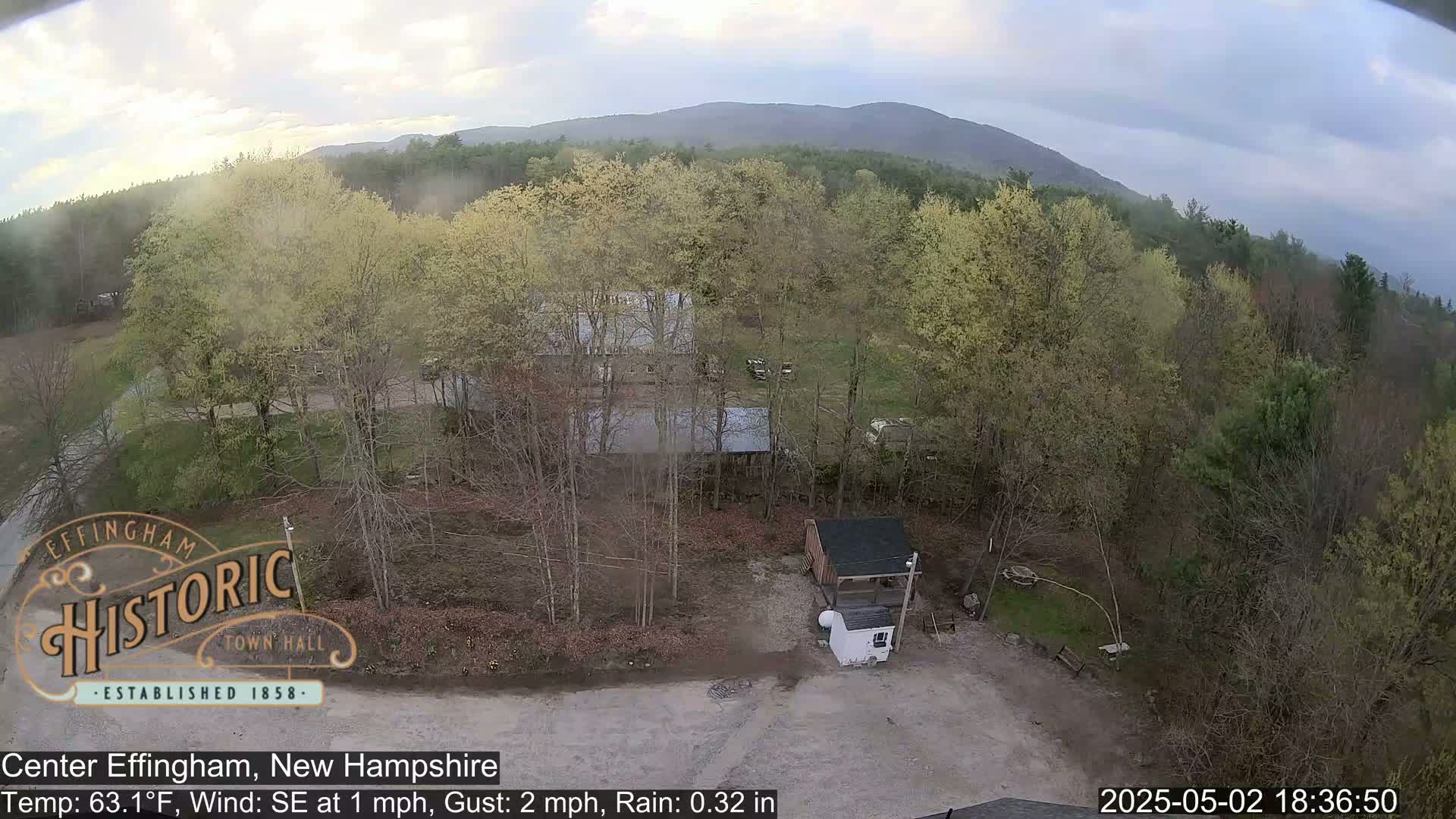 A high-angle view shows a lightly wooded area with several buildings, under an overcast sky with light rain.