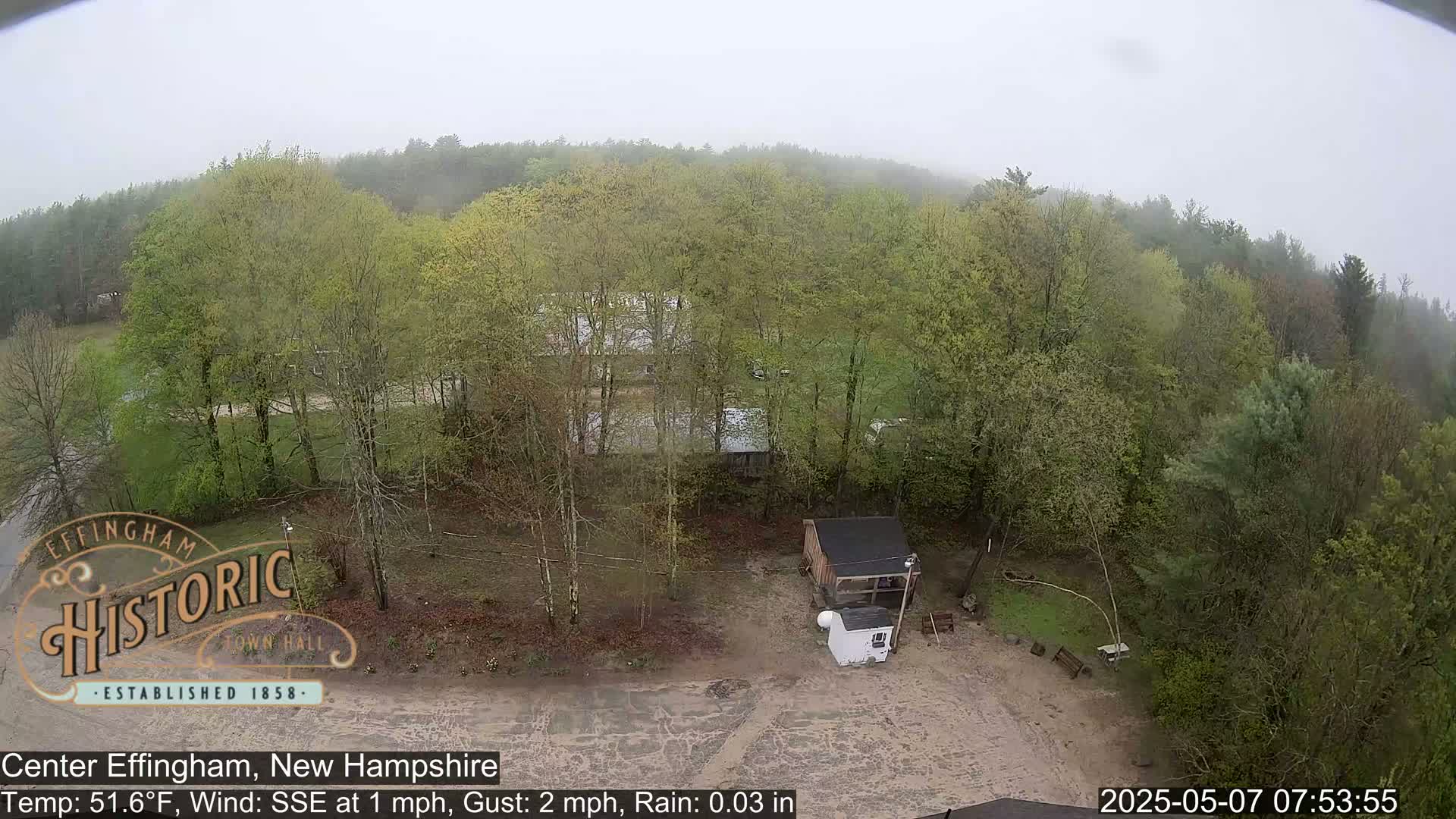 A high-angle view shows a small building in a lightly wooded area on a rainy day with light fog.