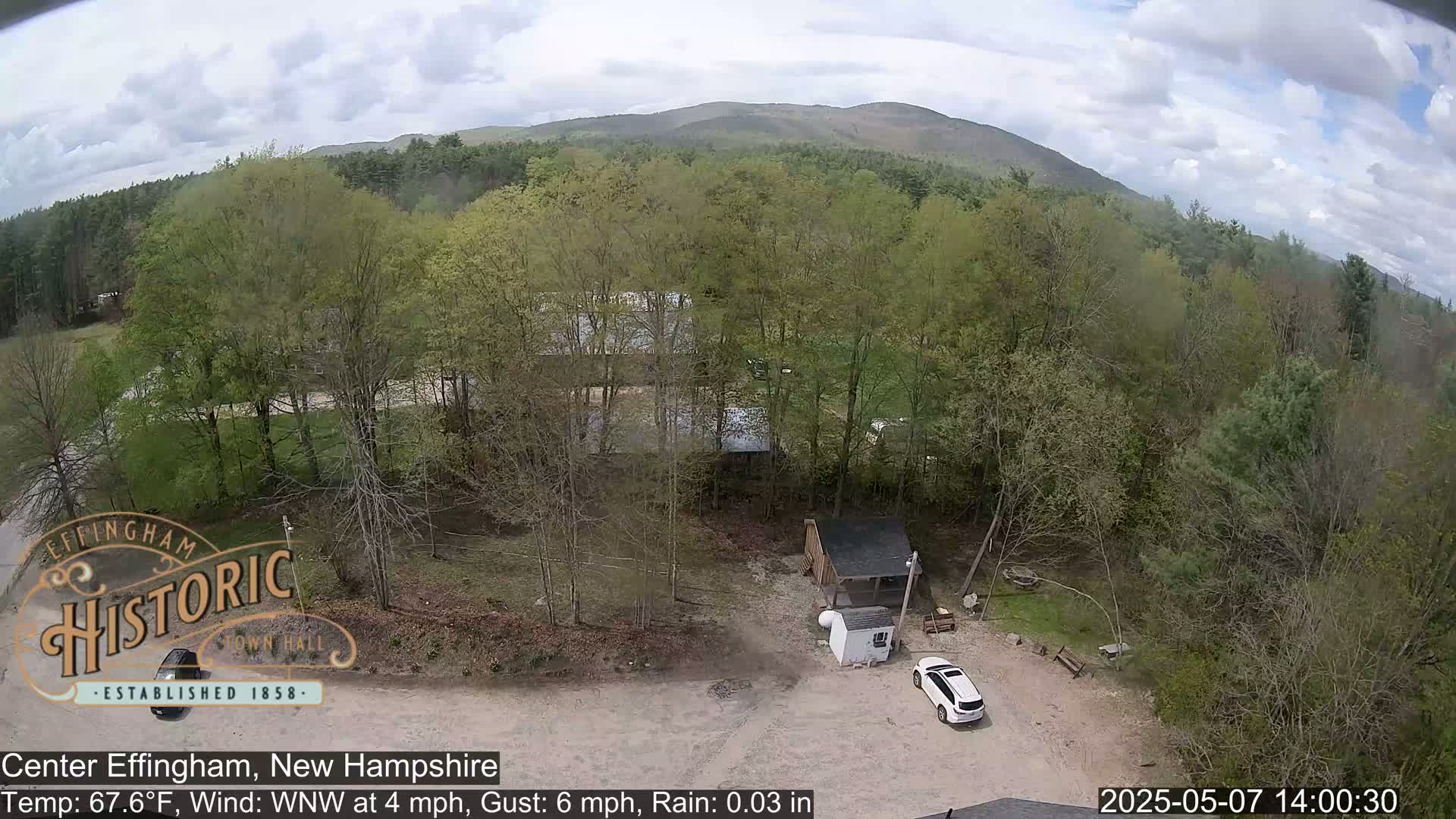 An aerial view shows a lightly wooded area with a small building, a white SUV, and a distant mountain range under partly cloudy skies with light rain.