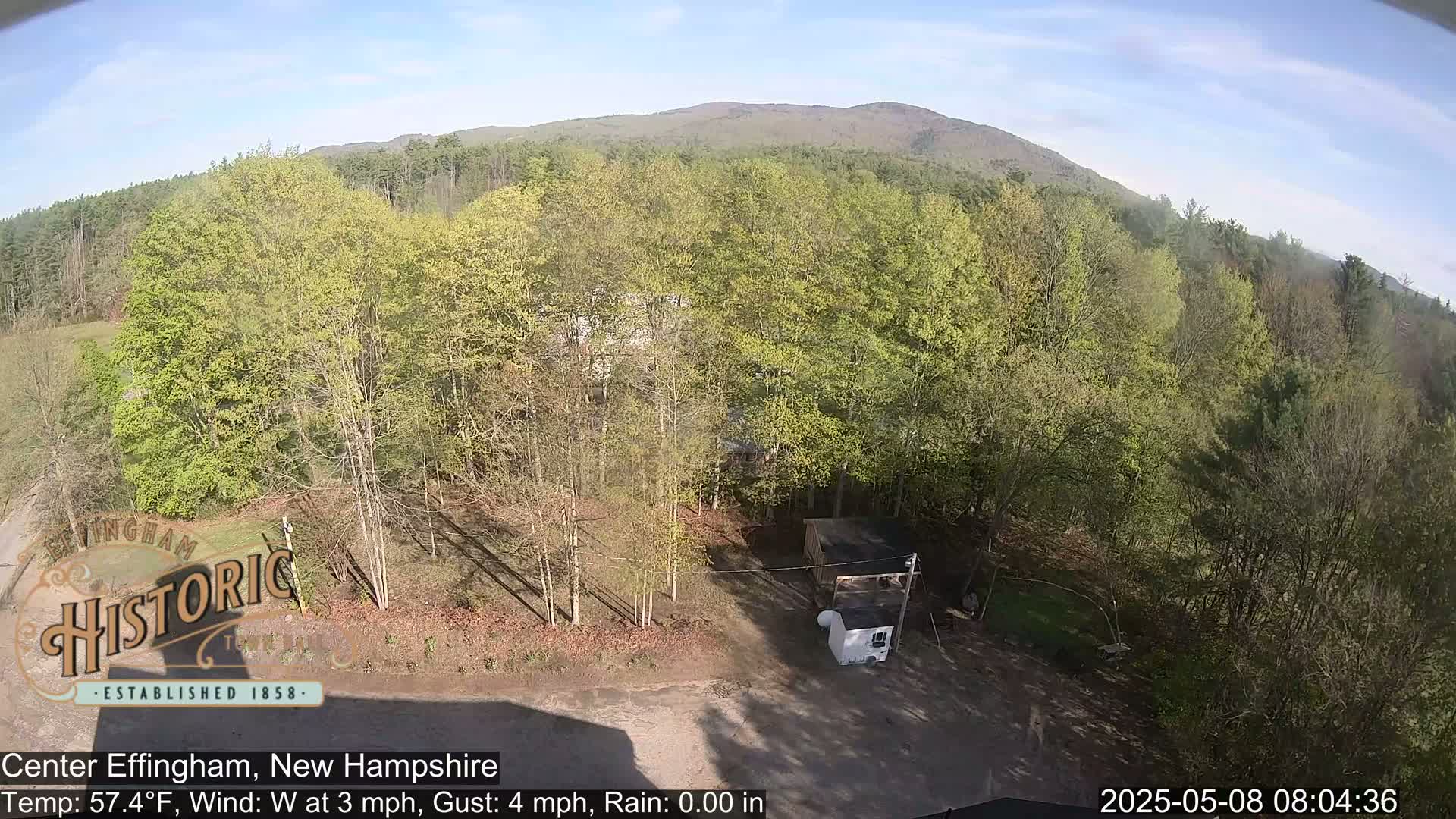 A high-angle view shows a mostly sunny day with a few light clouds over a wooded area, featuring some budding trees and a small shed in a clearing near the bottom of the frame, with a mountain visible in the distance.