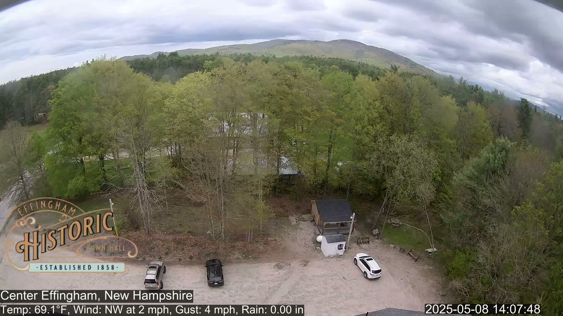 An aerial view shows a parking lot with two cars, a small shed, and lush green deciduous trees under a partly cloudy sky.