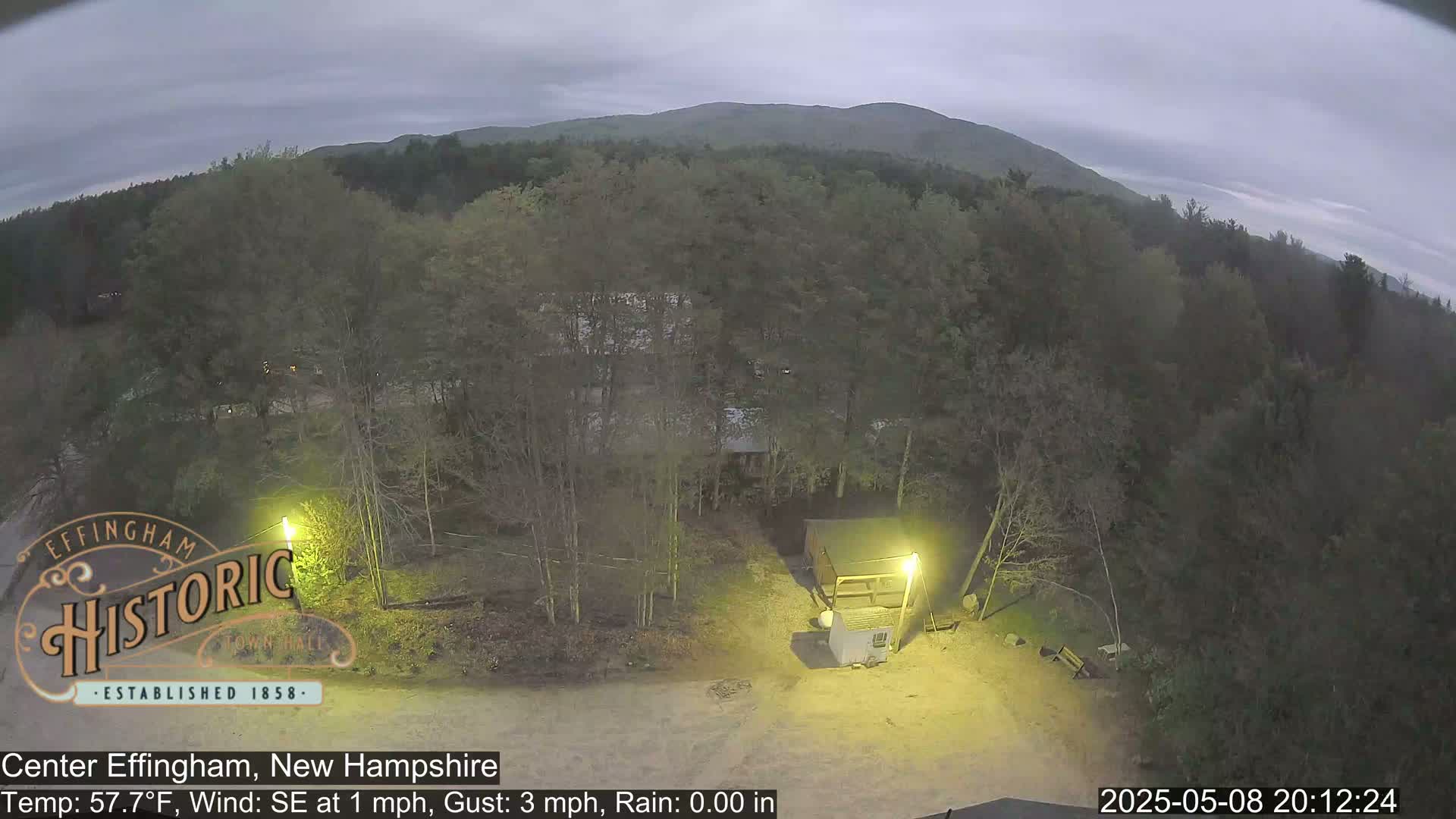 A nighttime, overcast view of a wooded area with a small building illuminated by a light, showing a calm scene with a slight southeastern wind.