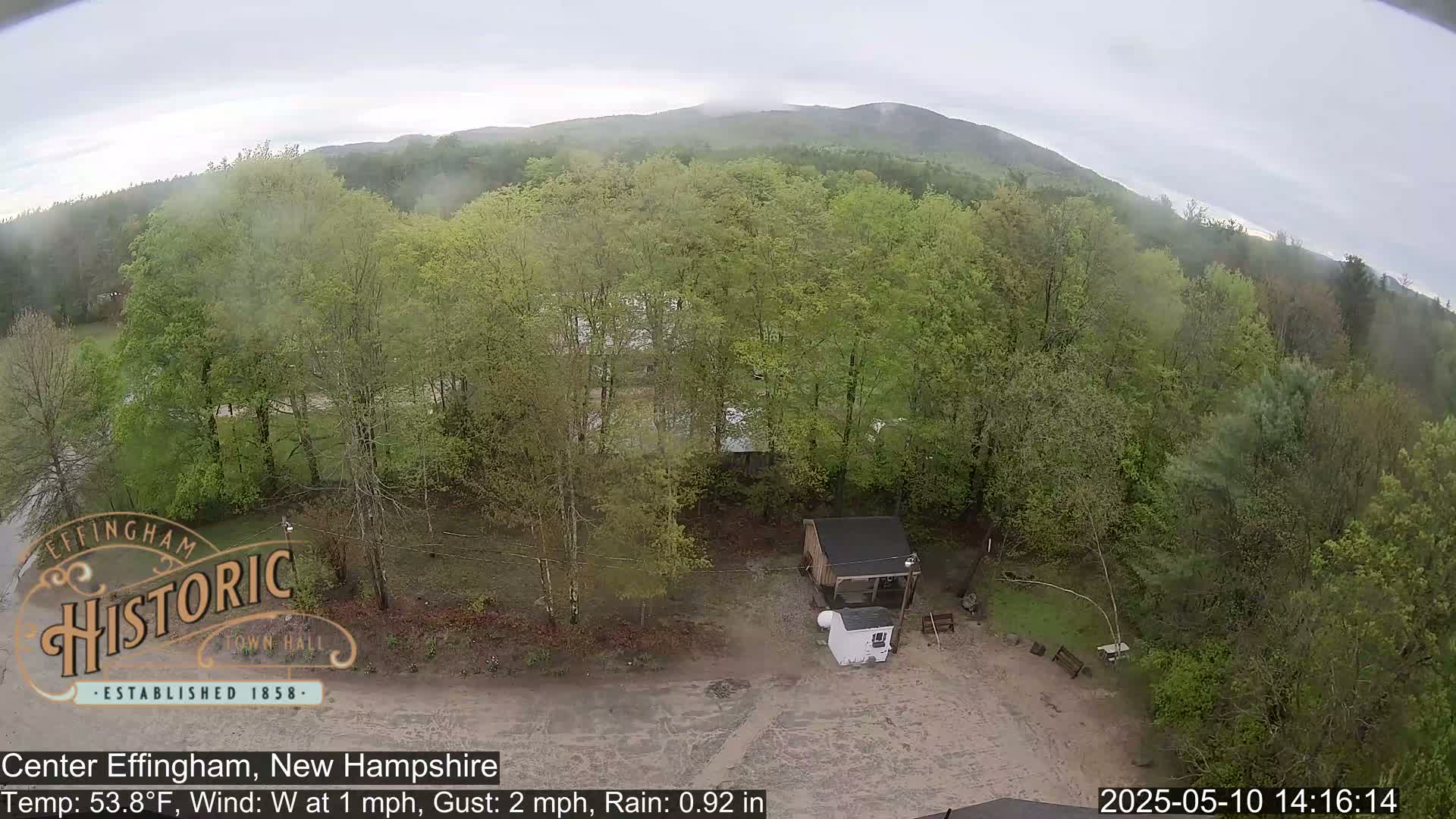 An aerial view shows a small building and a wooded area under a cloudy, rainy sky.