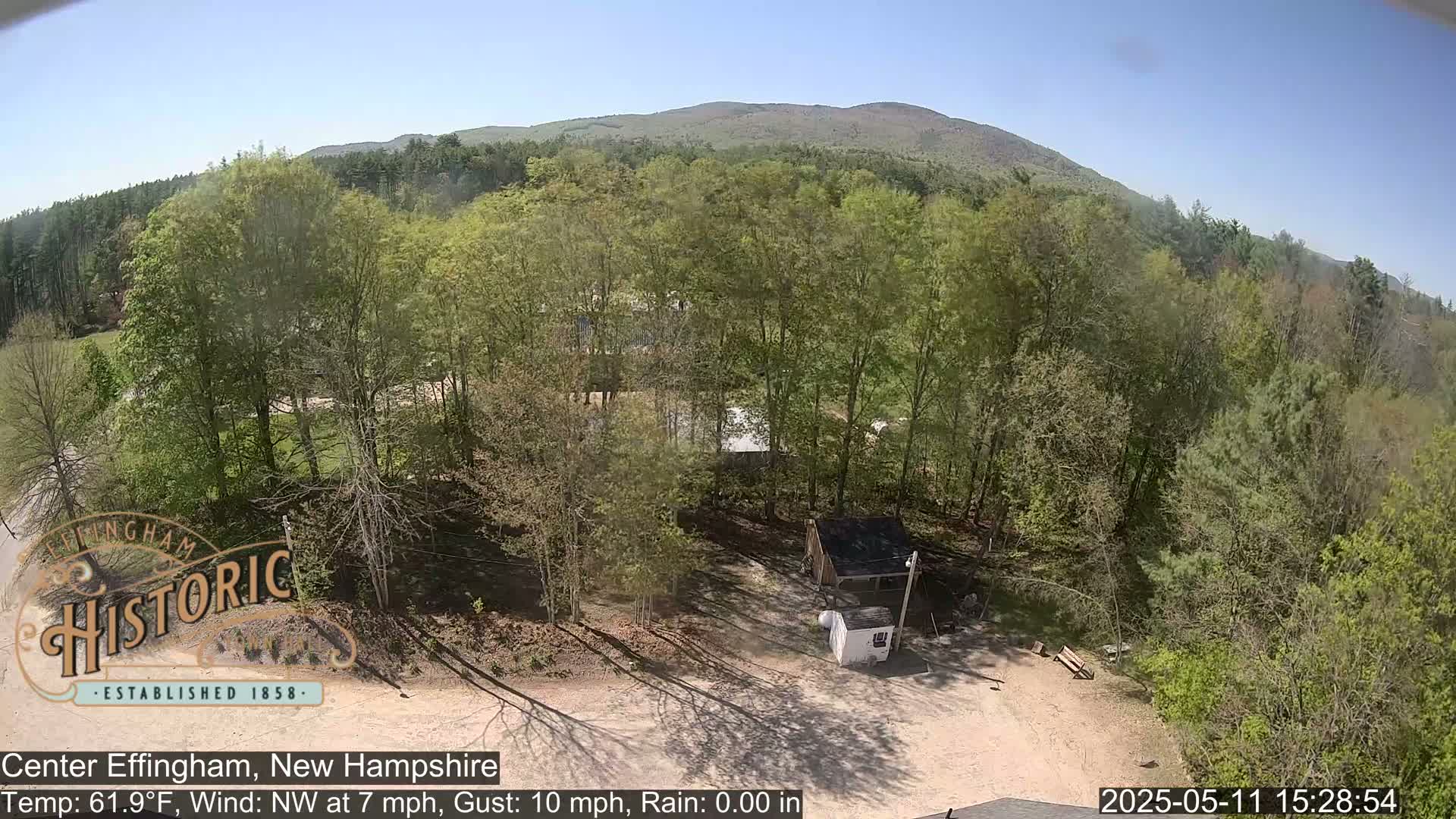 A high-angle view shows a small, wooded area with a small shed and a dirt parking lot, under a sunny sky with a mountain in the background.