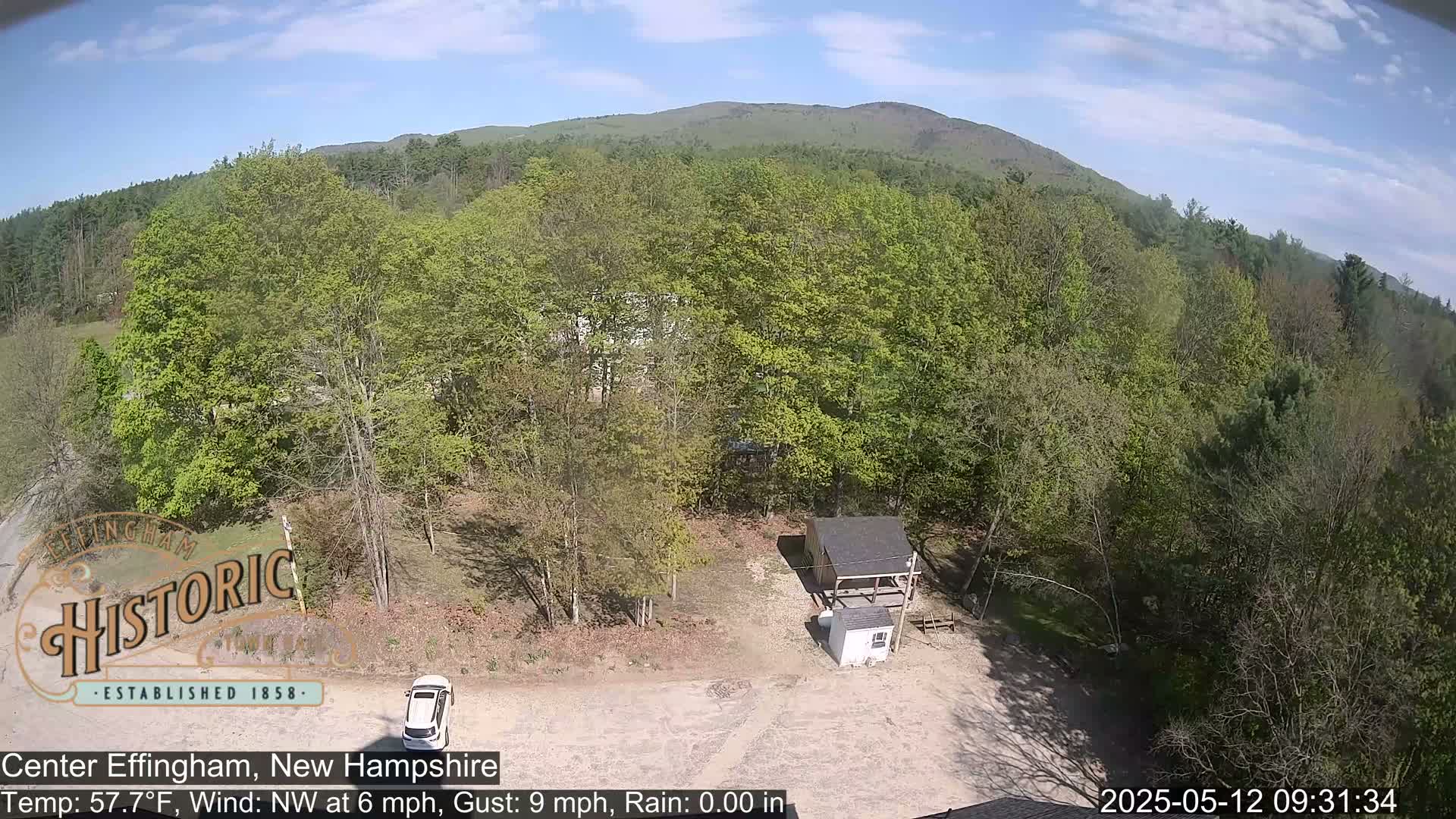 An aerial view shows a small building and a car in a clearing surrounded by lush green deciduous trees under a partly sunny sky.