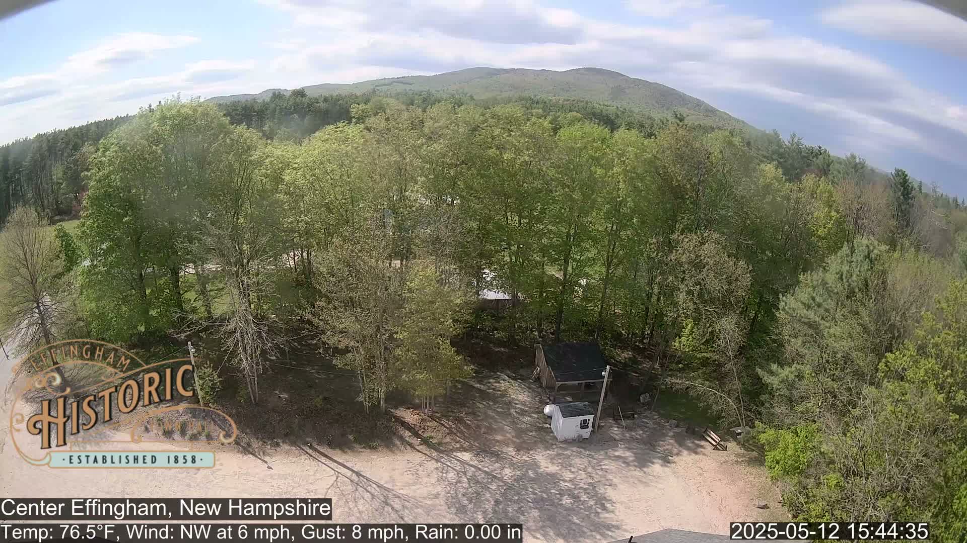A high-angle view shows a mostly sunny day over a woodland area with a small structure and a trailer visible in a clearing, near a small road, with a hill in the distance.