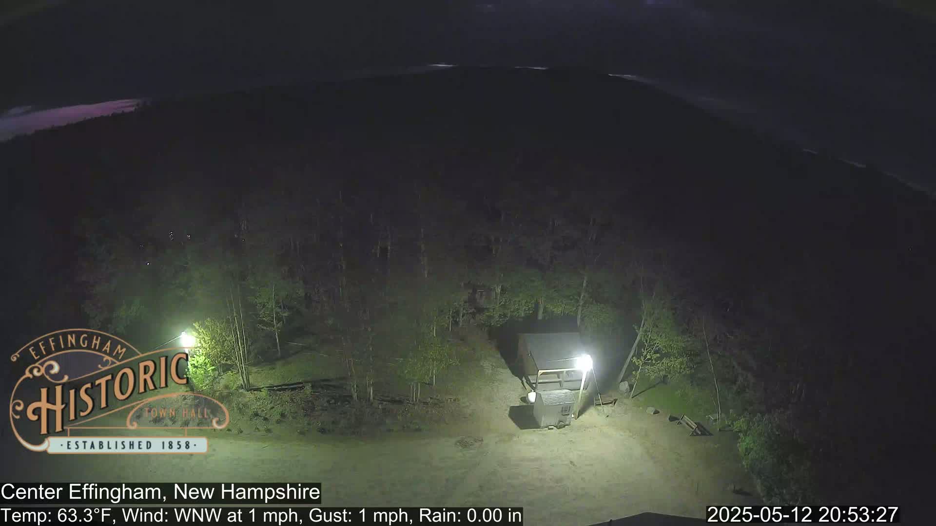 A nighttime, overhead view shows a small building in a wooded area under a slightly cloudy sky with light rain.