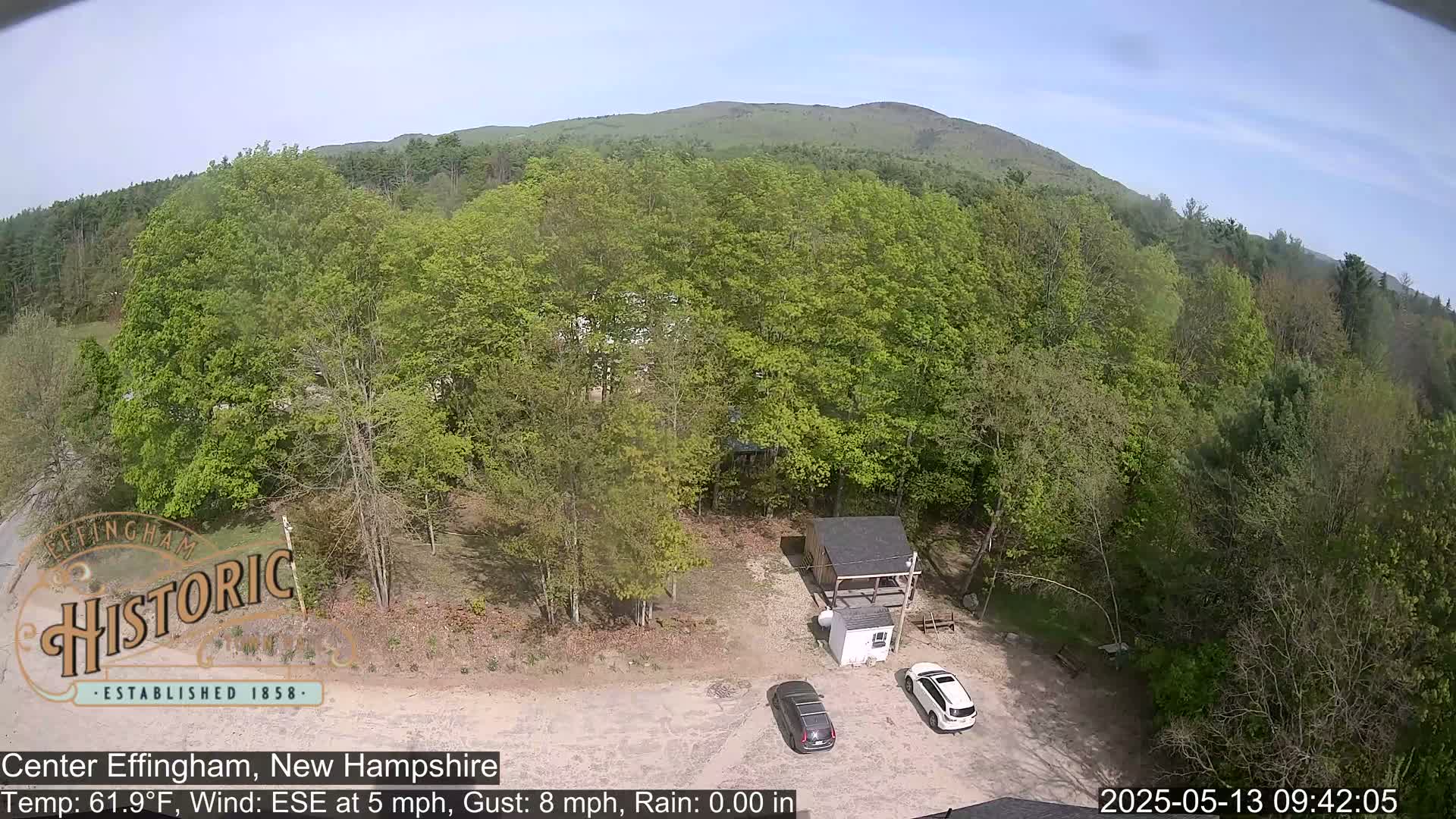 An aerial view shows a parking lot with two cars next to a small building surrounded by lush green trees and a distant mountain under a clear sky.