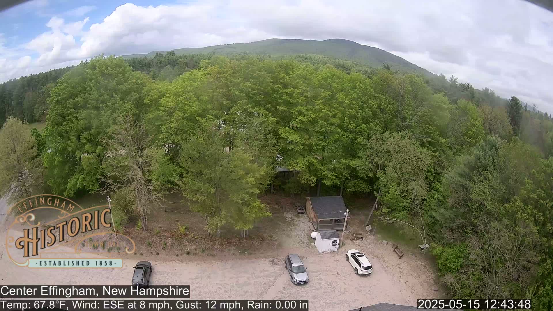 An aerial view shows a small parking lot with two cars, a small building nestled within a lush green forest, and a mountain range in the background under partly cloudy skies.