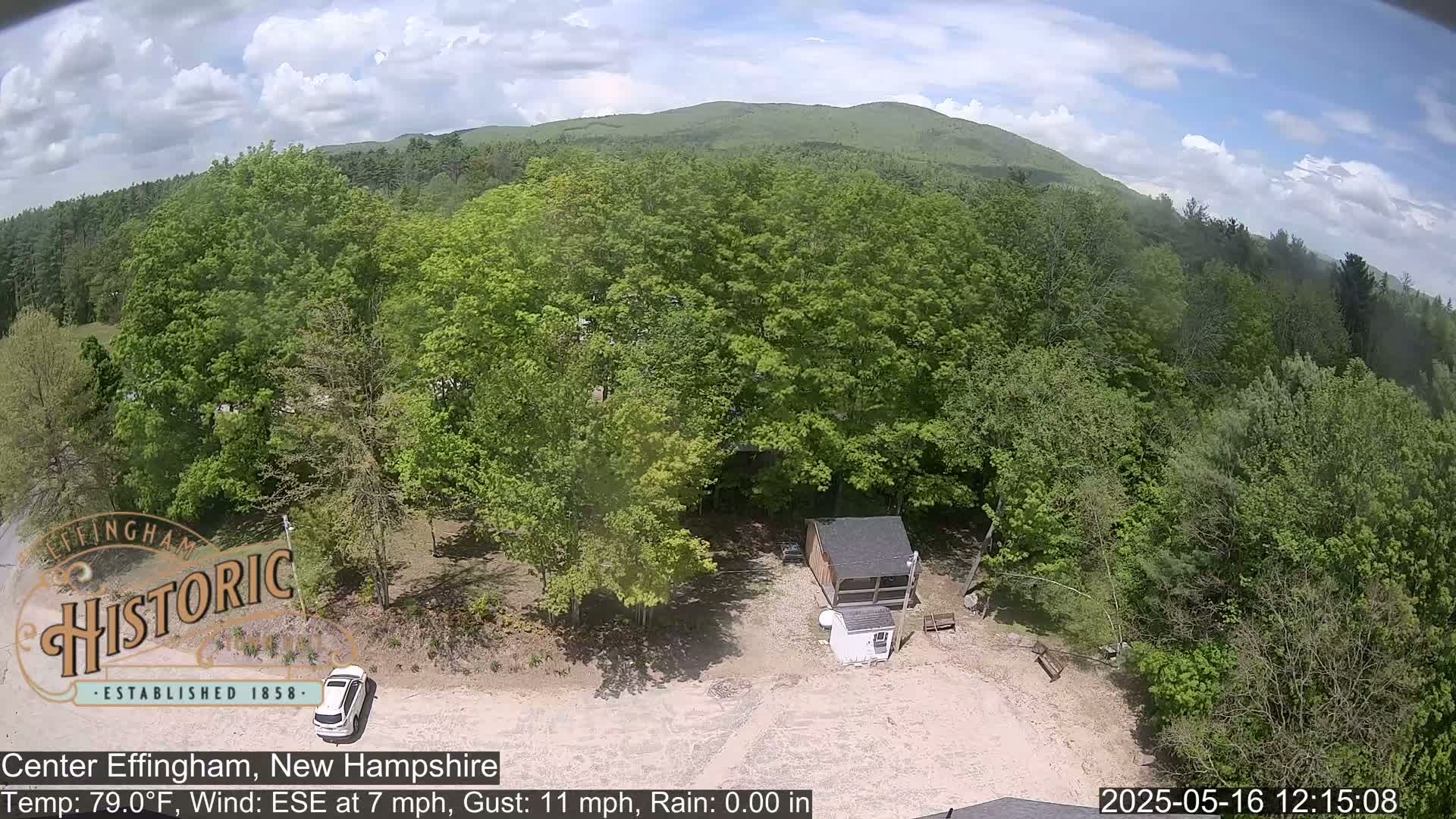 An aerial view shows a small cabin nestled among lush green trees on a partly cloudy day, with a car parked nearby.