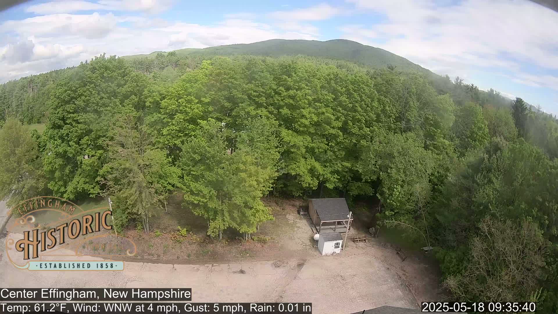 An aerial view shows a small shed and a mostly-wooded area with light rain under a partly cloudy sky.