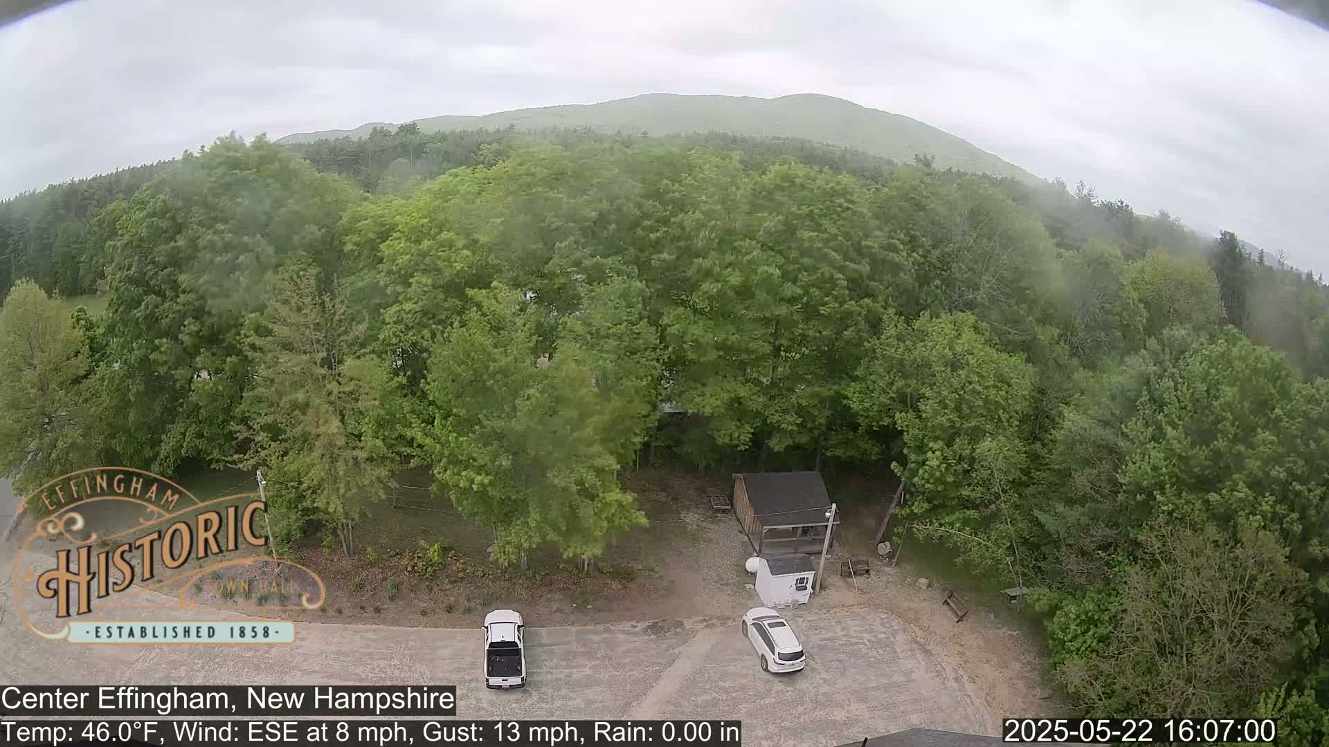 An aerial view shows a parking lot with two cars near a small shed surrounded by lush green trees and a distant mountain under an overcast sky.