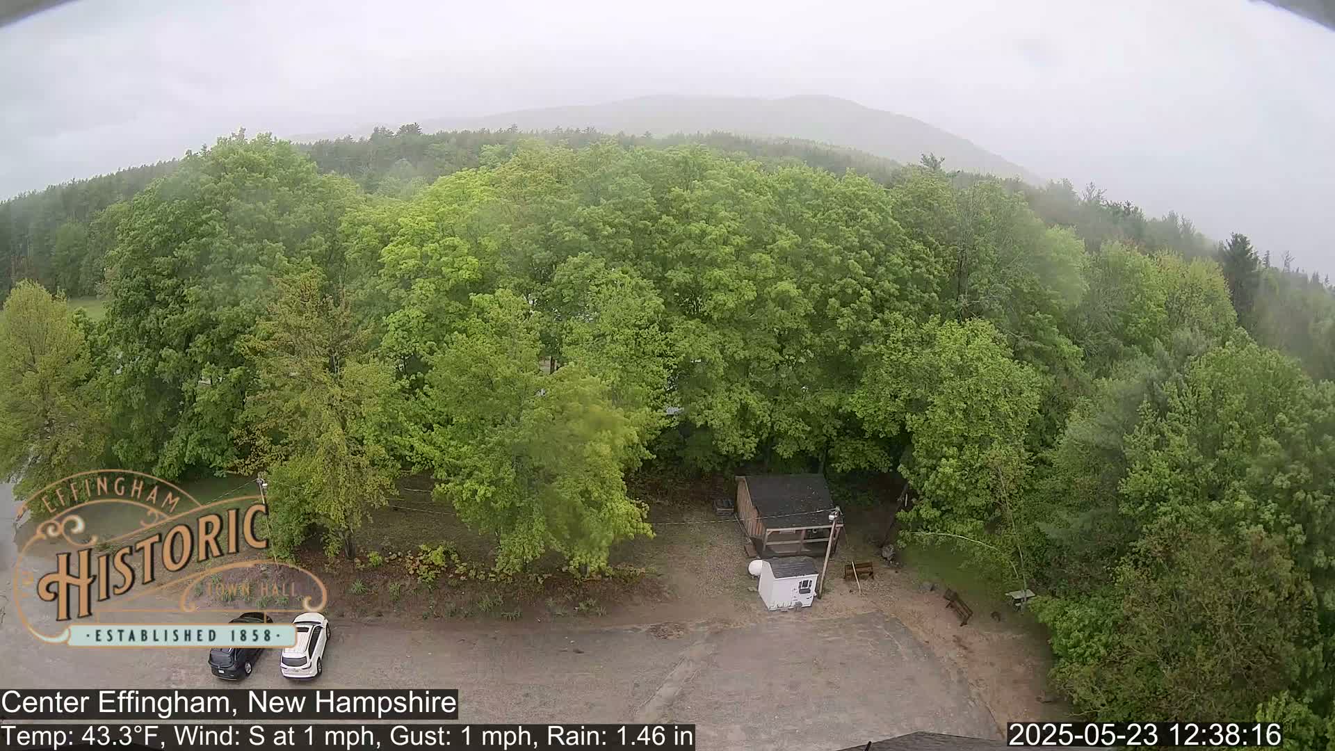 An aerial view shows a small building and a parking lot surrounded by lush green trees under a rainy sky.