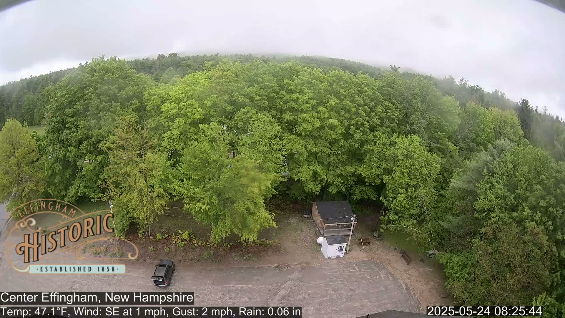 An aerial view shows a parking lot, a small building, and lush green trees under a foggy, rainy sky.