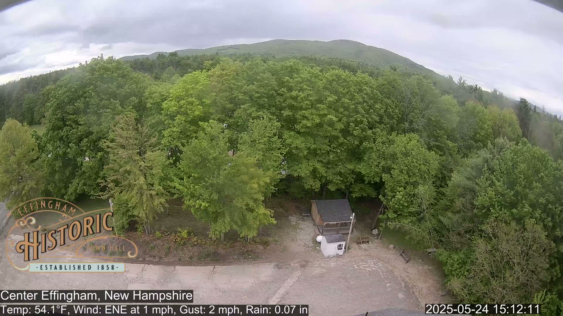 An aerial view shows a small building surrounded by lush green trees under a gray, overcast sky with light rain.