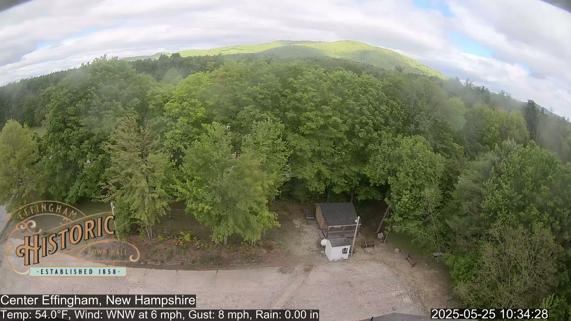 An elevated view shows a small shed in a clearing surrounded by lush green deciduous trees, with distant mountains visible under a partly cloudy sky.