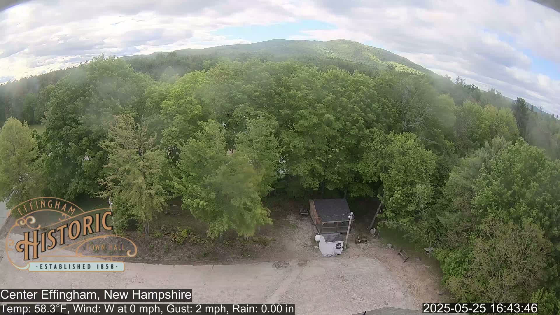 A high-angle view shows a small building and a mostly-wooded area under a cloudy sky with light rain.