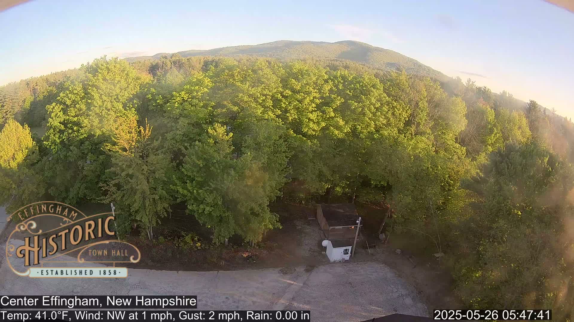 An elevated view shows a mostly sunny area with lush green trees, a small shed, and a mountain in the distance, under calm conditions.