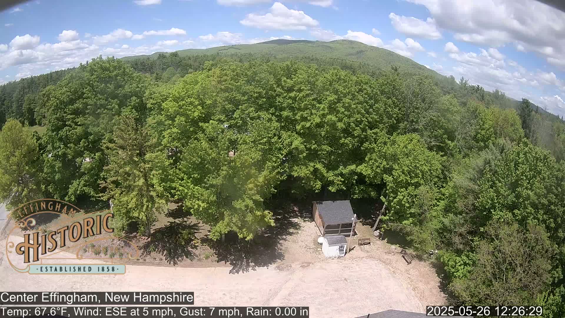 An aerial view shows a small building in a clearing surrounded by lush green deciduous trees under a partly cloudy sky.