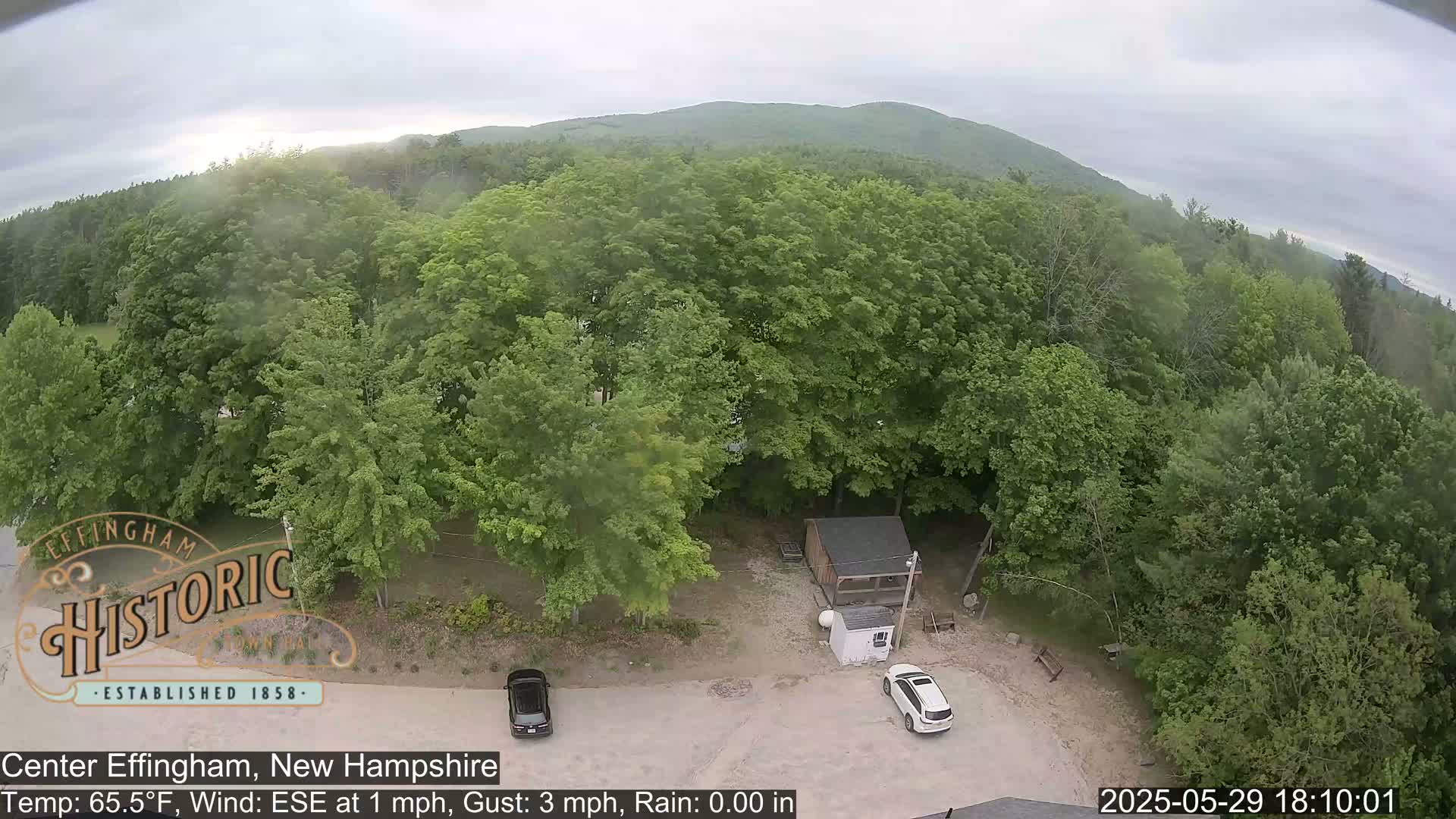 An aerial view shows a small parking lot with two cars, a small shed, and lush green trees surrounding a clearing under an overcast sky.