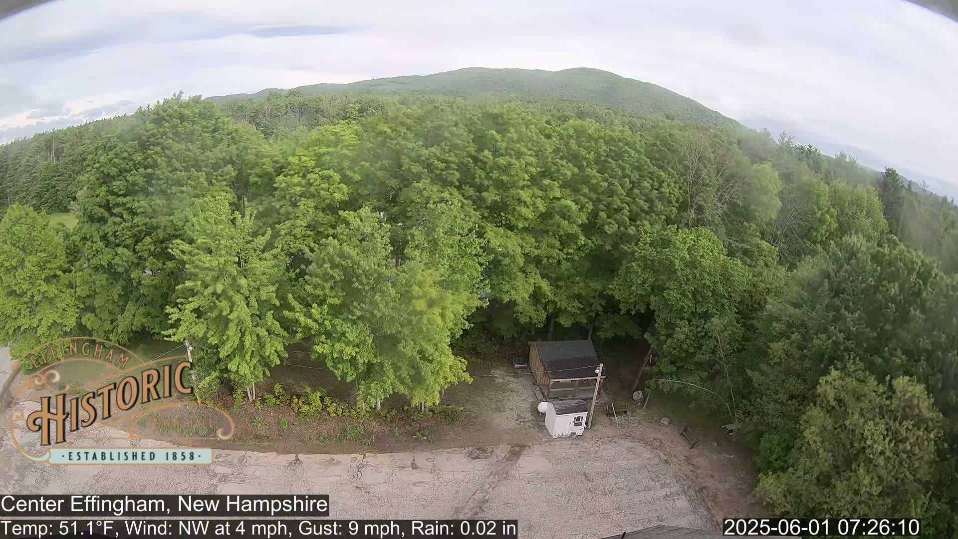 An aerial view shows a small building in a clearing surrounded by lush green trees under a cloudy sky with light rain.