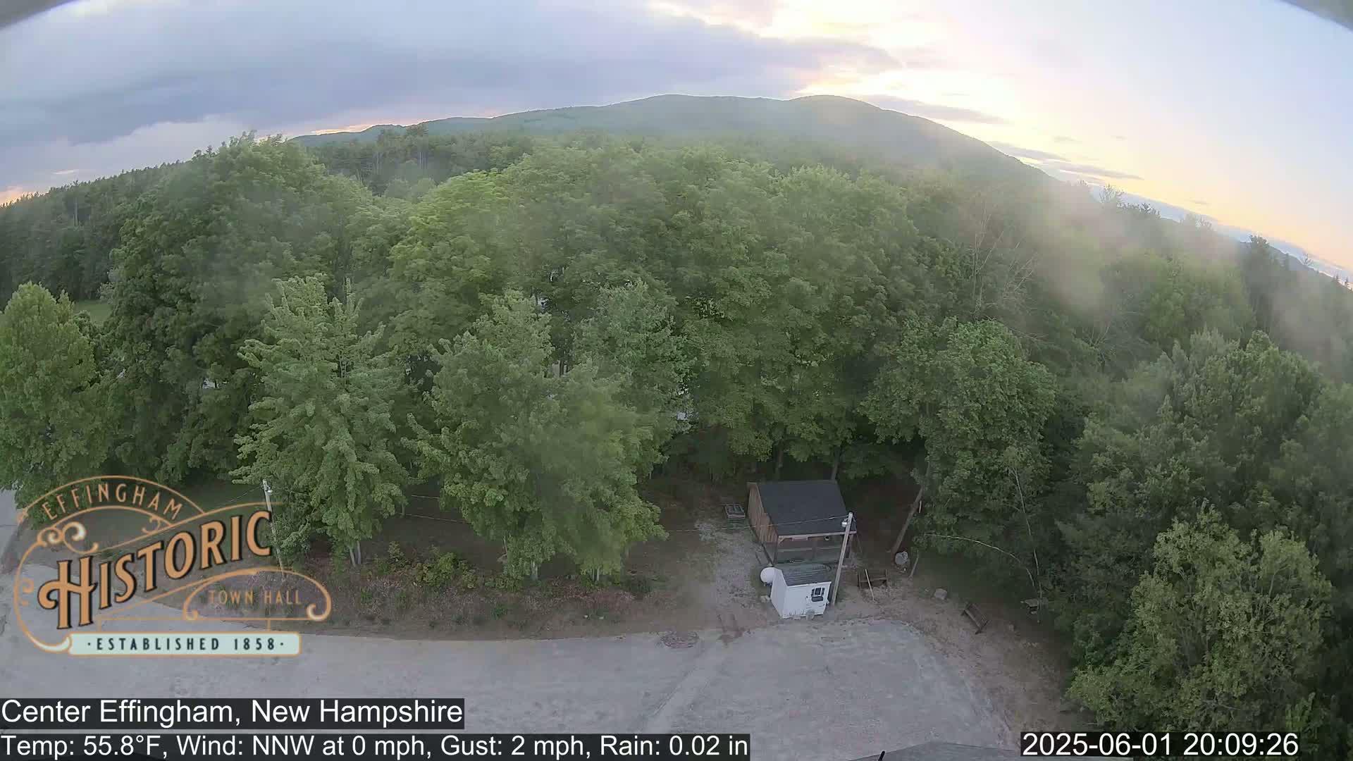An aerial view shows a light rain falling over a verdant forest with a small building and a gravel area in the foreground, and mountains in the background under a partly cloudy sky.