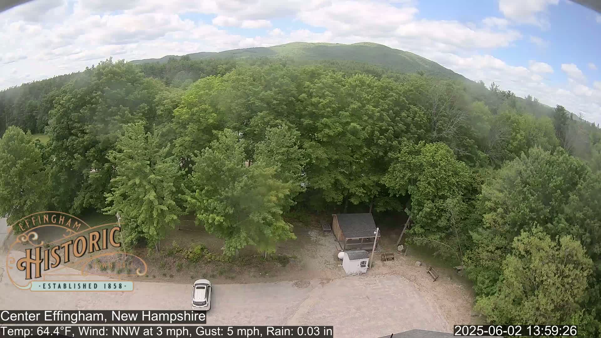 An aerial view shows a lightly wooded area with a small building and a car parked in a lot, under a partly cloudy sky with light rain.