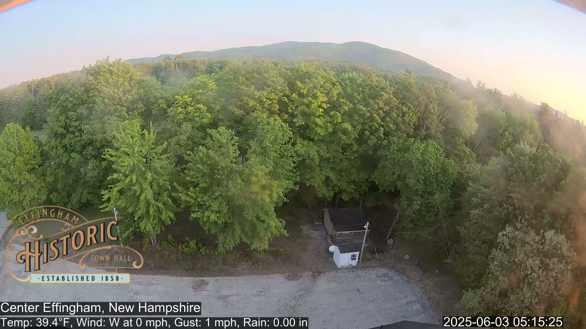 An aerial view shows a small building surrounded by lush green trees under a partly hazy sky.