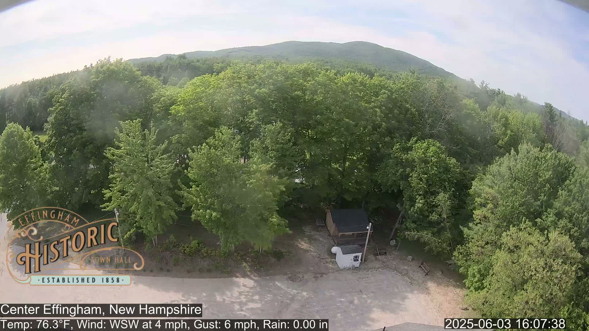 An aerial view shows a small building and a camper in a clearing surrounded by lush green deciduous trees and a distant mountain under partly cloudy skies.