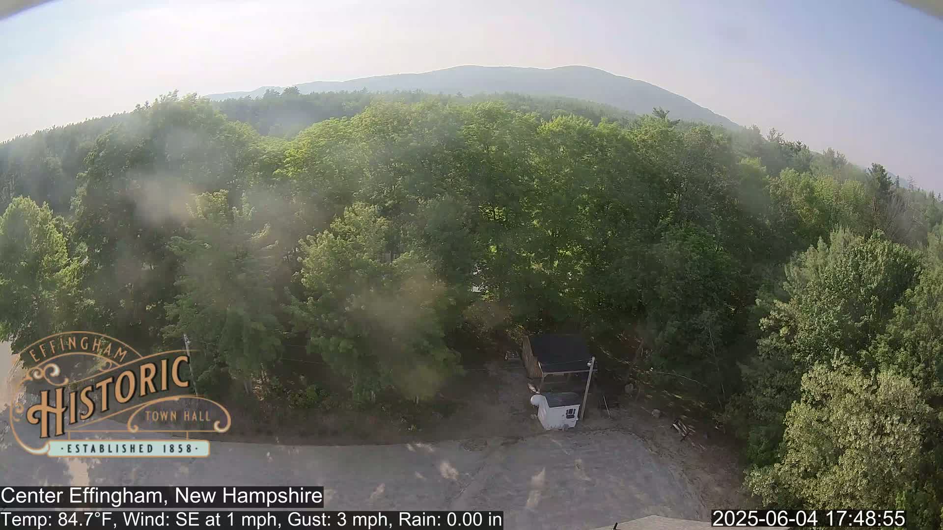 An elevated view shows a hazy, sunny day overlooking a lush green forest with a small structure and a paved area below, mountains in the distance.