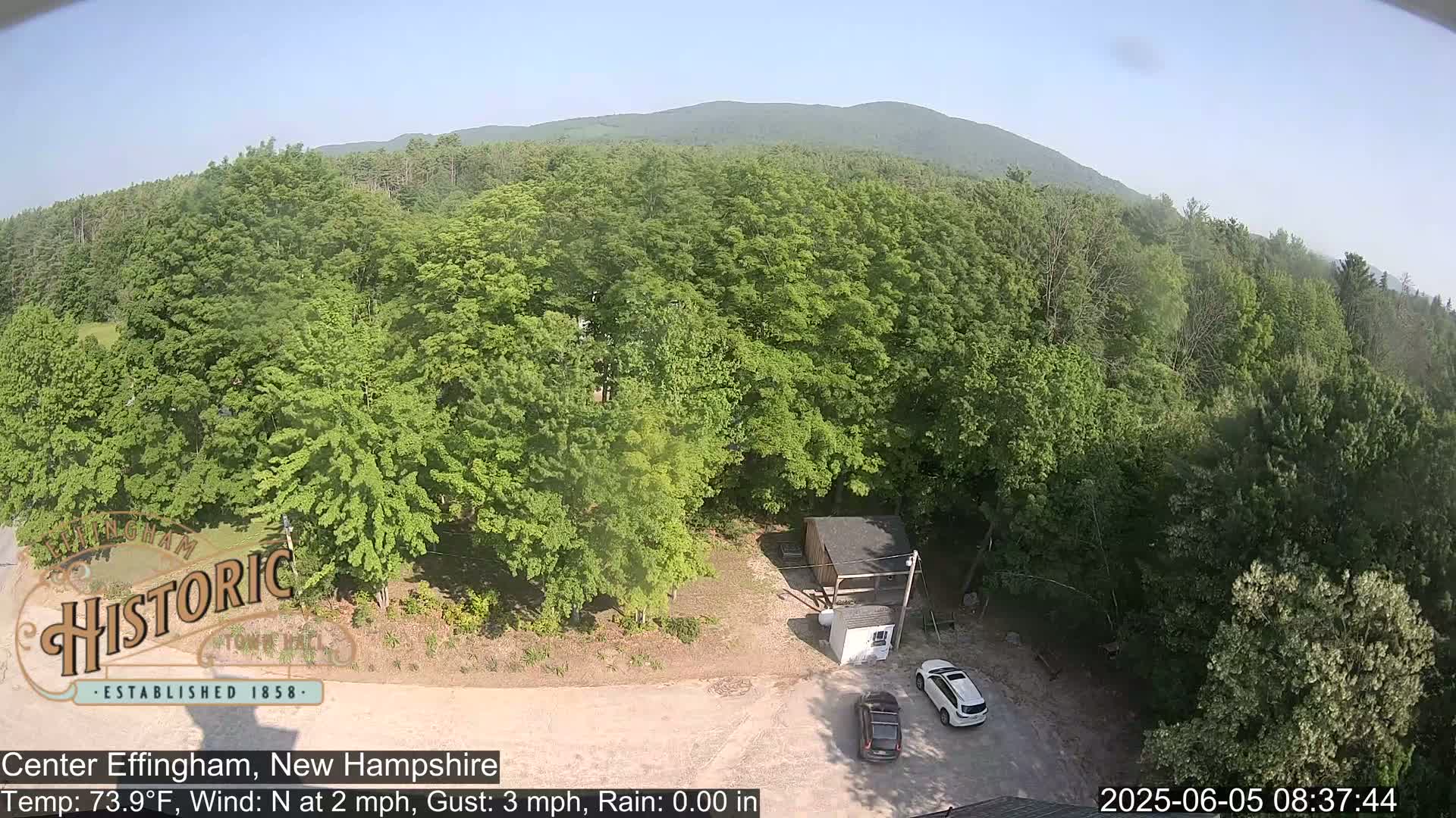 An aerial view shows a parking lot with two cars near a small building surrounded by lush green trees and a distant mountain under a clear sky.