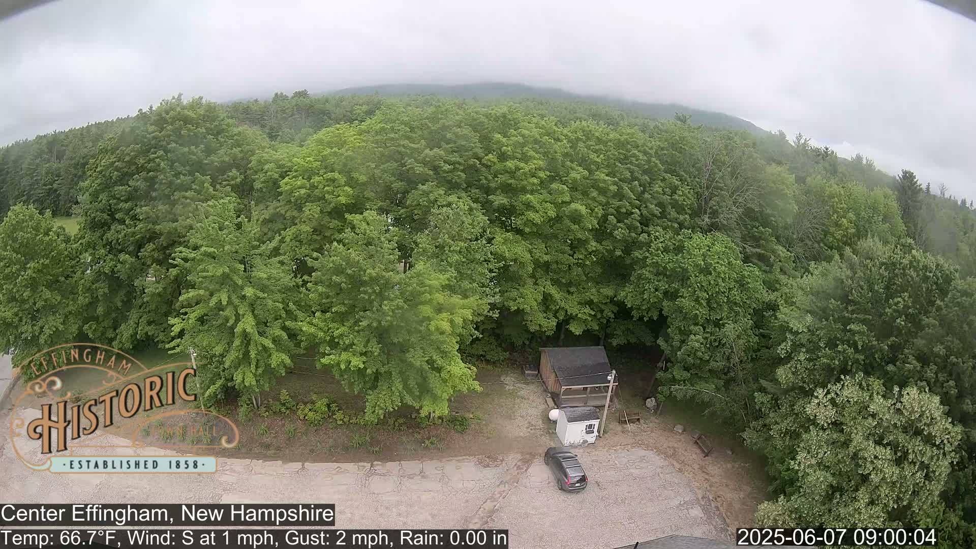 A dark-colored SUV is parked in a small lot next to a small building surrounded by lush green trees under an overcast sky.