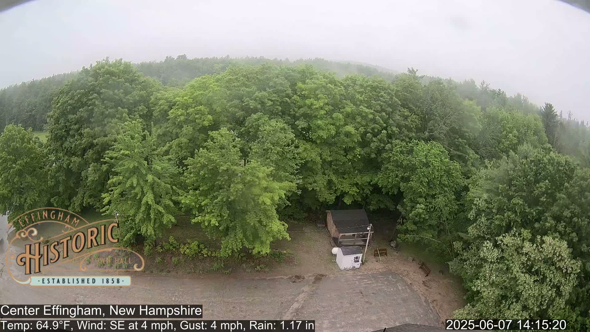 A high-angle view shows a small shed in a clearing surrounded by lush green trees under rainy conditions.