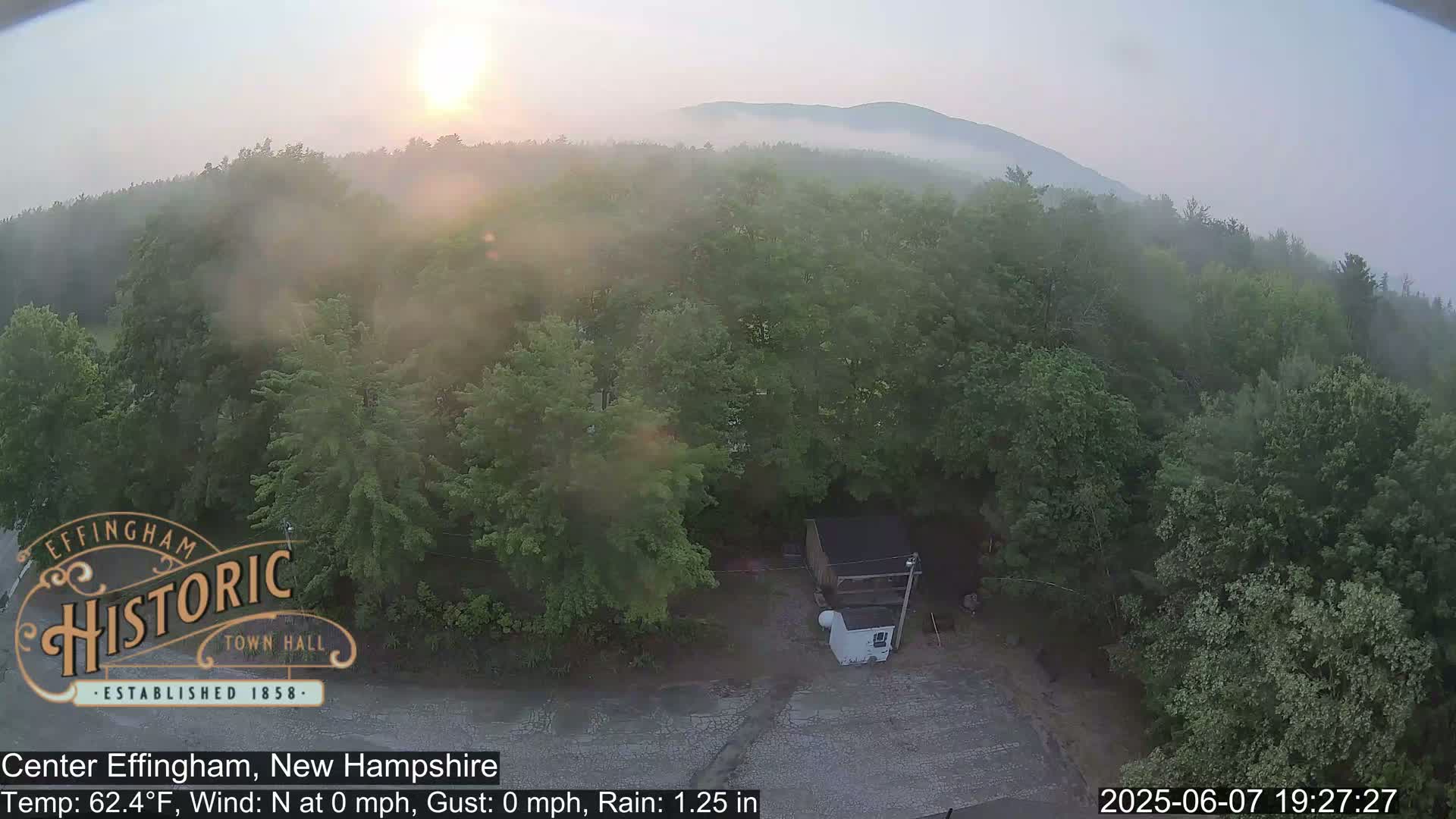 A misty sunrise illuminates a forested landscape with a small structure visible in the foreground, under rainy conditions.