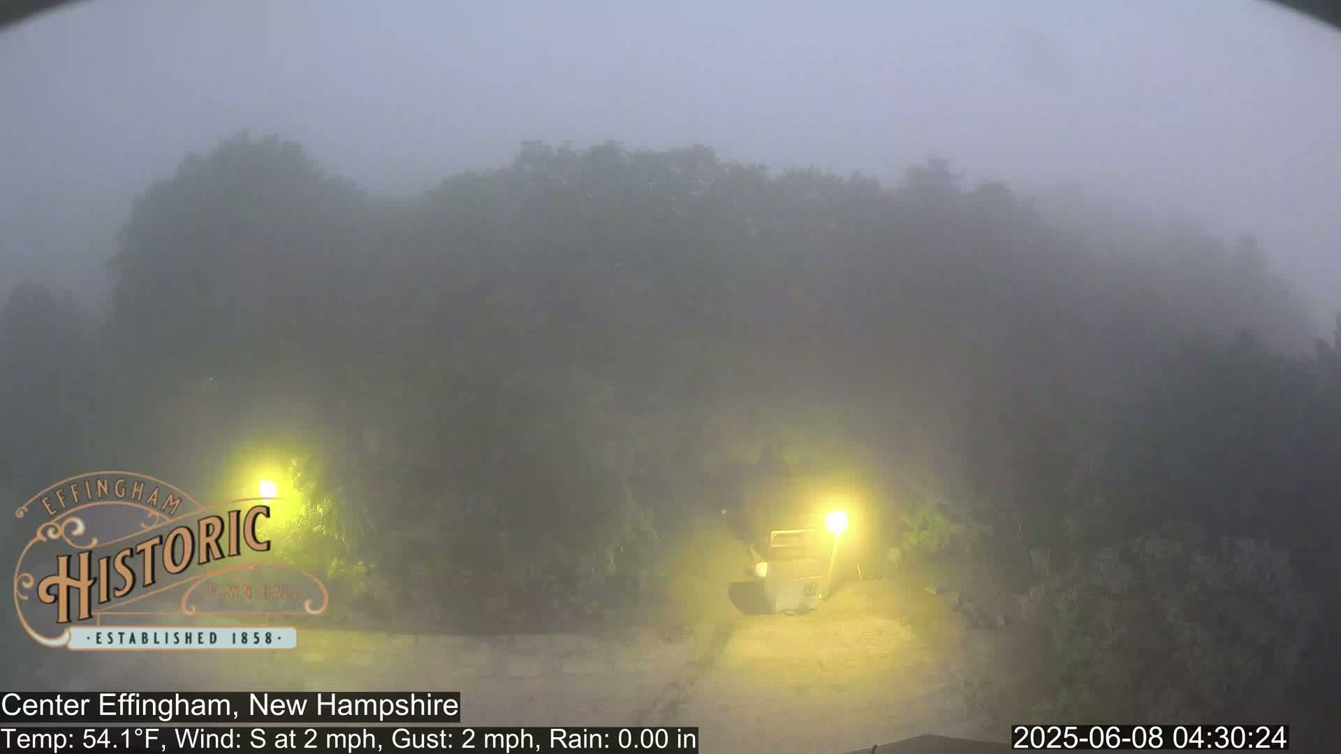 A foggy scene shows a road with a building and trees shrouded in mist under lightly lit conditions.