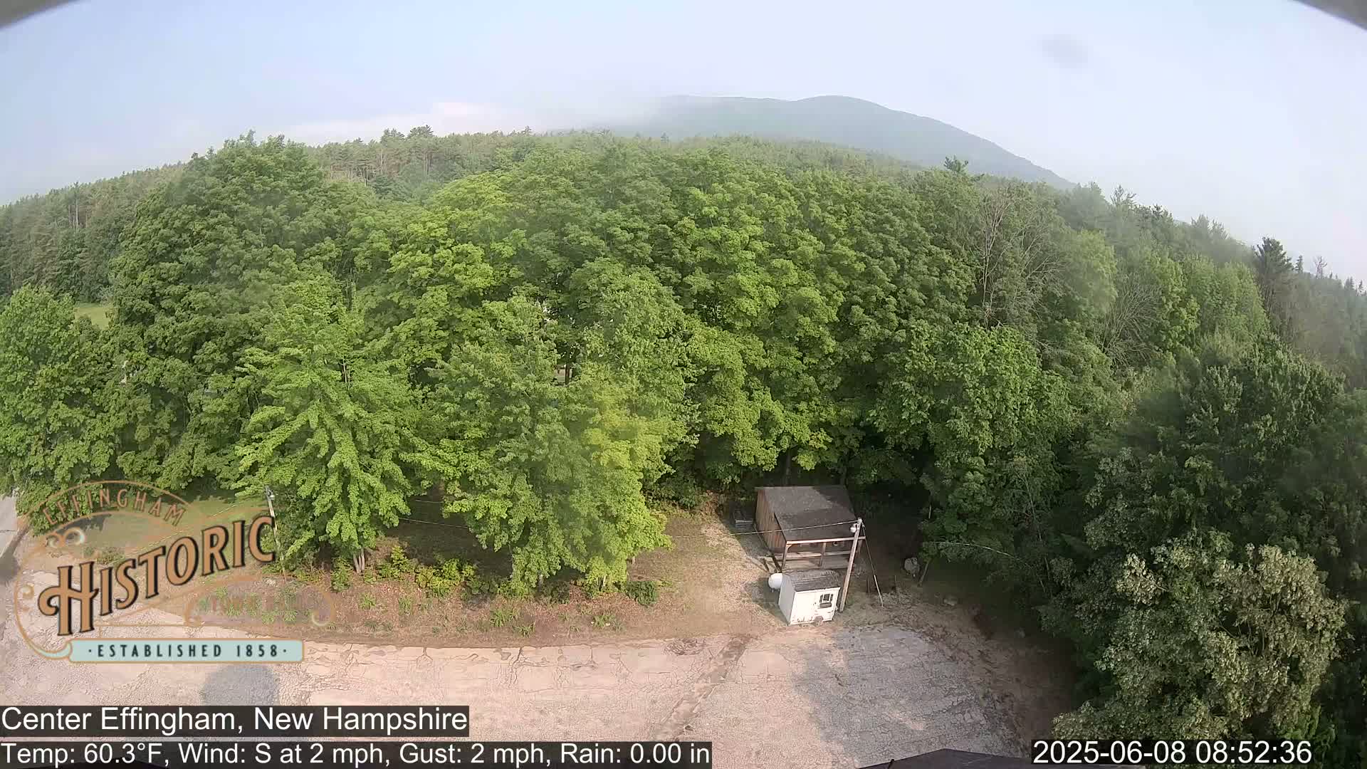 A high-angle view shows a mostly clear day over a wooded area with a small shed and a white structure in a gravel area; a hazy mountain is visible in the background.