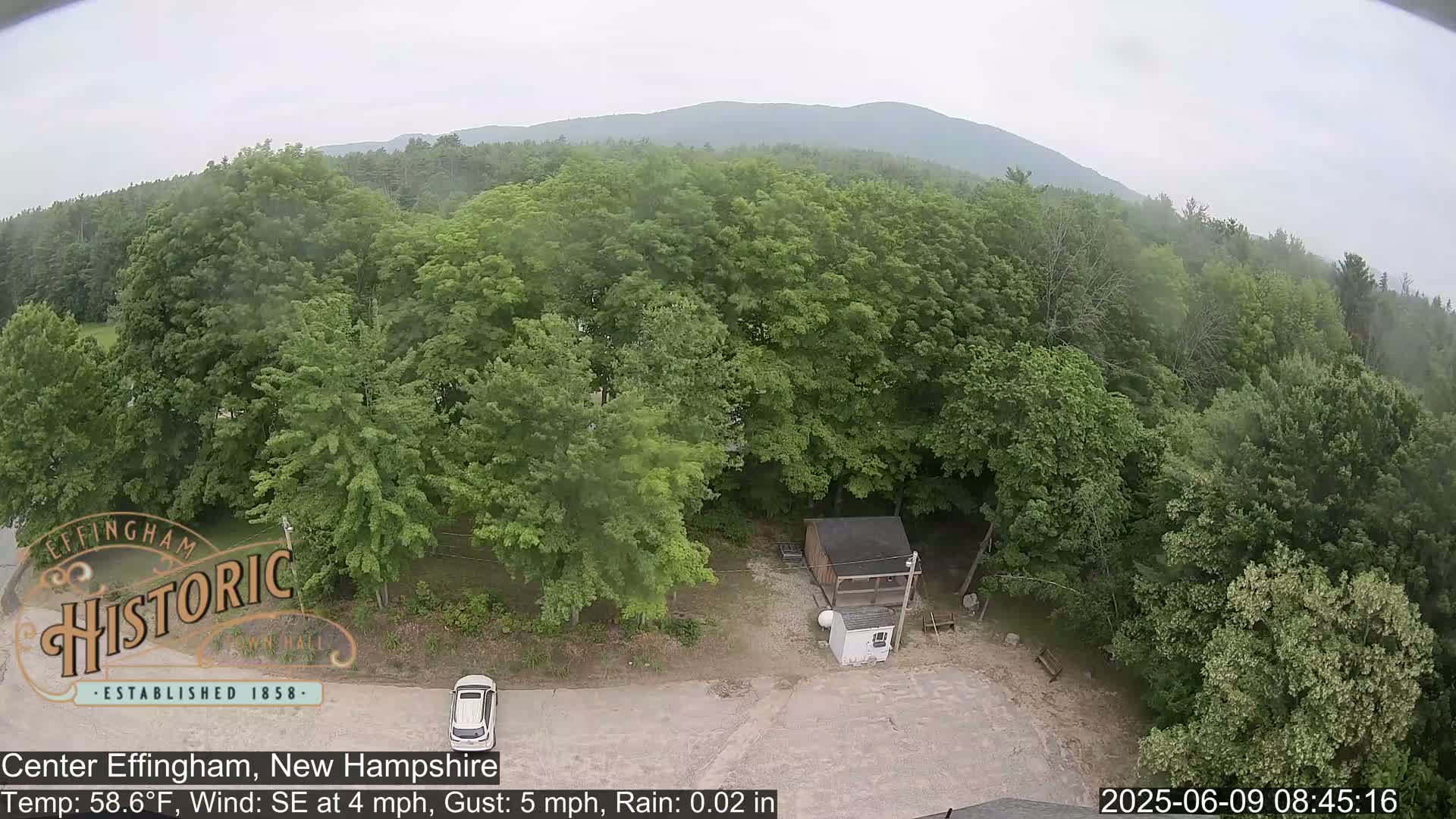A high-angle view shows a light rain falling on a parking lot next to a wooded area with a small building and a car, all situated in front of a mountain range under an overcast sky.